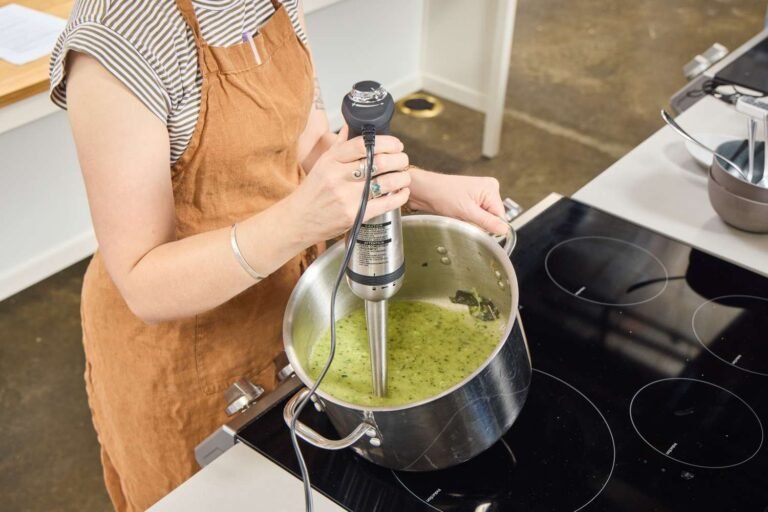 An immersion blender blending bright orange carrot soup in a glass pitcher.