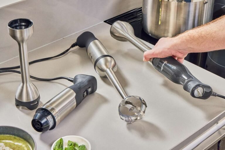 Stainless steel immersion blender beside a bowl of smooth carrot soup on a marble counter.